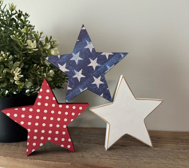 May include: Three wooden stars in red, white, and blue patriotic colors. The red star has white polka dots, the blue star has white stars, and the white star has a gold outline.