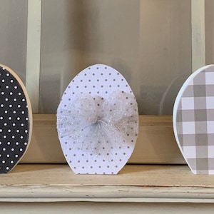 May include: Five wooden eggs decorated with different patterns, including black and white gingham, black and white polka dots, gray and white gingham, and gray and white polka dots. Each egg has a black tulle bow.