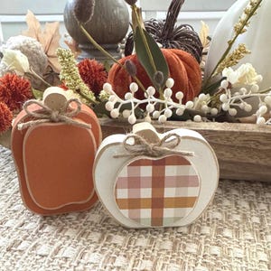 May include: Two wooden pumpkin-shaped decorations. The left pumpkin is solid orange with a natural wood stem and a jute twine bow. The right pumpkin is white with a central plaid pattern in shades of orange, brown, and cream, featuring a jute twine bow.
