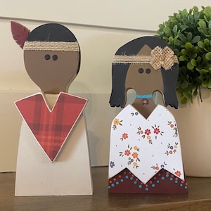 May include: Two wooden figurines of Native American people. The male figurine is wearing a red and black plaid shirt and a brown headband. The female figurine is wearing a white dress with a floral pattern and a brown headband with a brown flower.