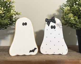 Puede incluir: Dos decoraciones de fantasmas de madera blanca. Un fantasma tiene un murciélago negro, el otro tiene un lazo de lunares blanco y negro. Ambos tienen ojos ovalados negros. Los fantasmas están sobre una superficie de madera con plantas verdes en macetas negras a cada lado.