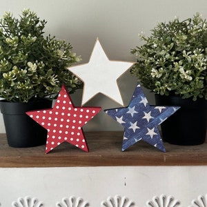 May include: Three wooden stars, each with a different design. The first star is white, the second is red with white polka dots, and the third is blue with white stars. The stars are sitting on a wooden shelf in front of two potted plants.