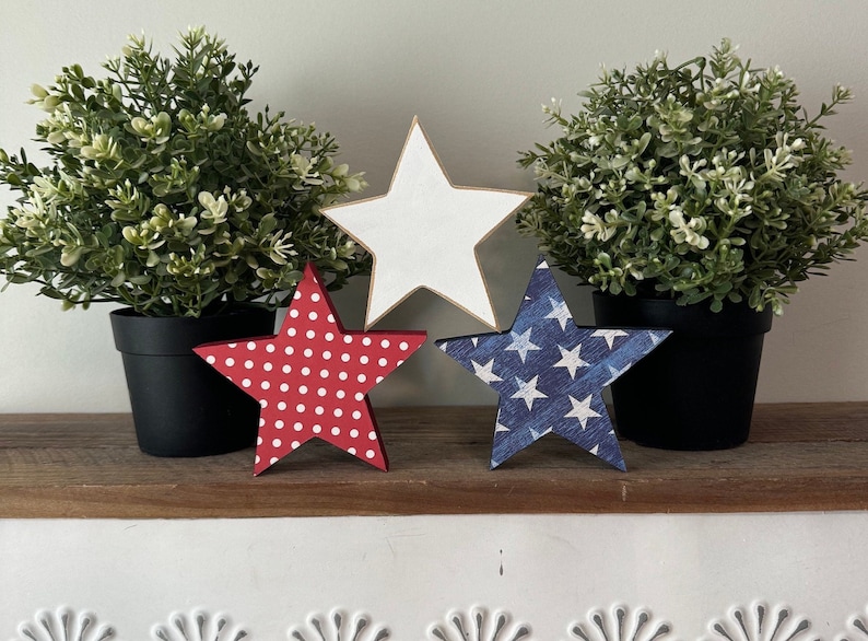 May include: Three wooden stars, each with a different design. The first star is white, the second is red with white polka dots, and the third is blue with white stars. The stars are sitting on a wooden shelf in front of two potted plants.
