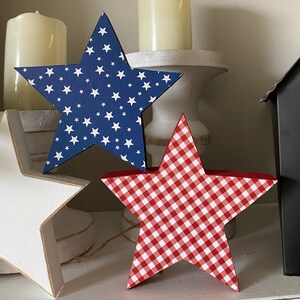 May include: Two wooden stars, one blue with white stars and one red with white gingham pattern. The stars are sitting on a white surface.