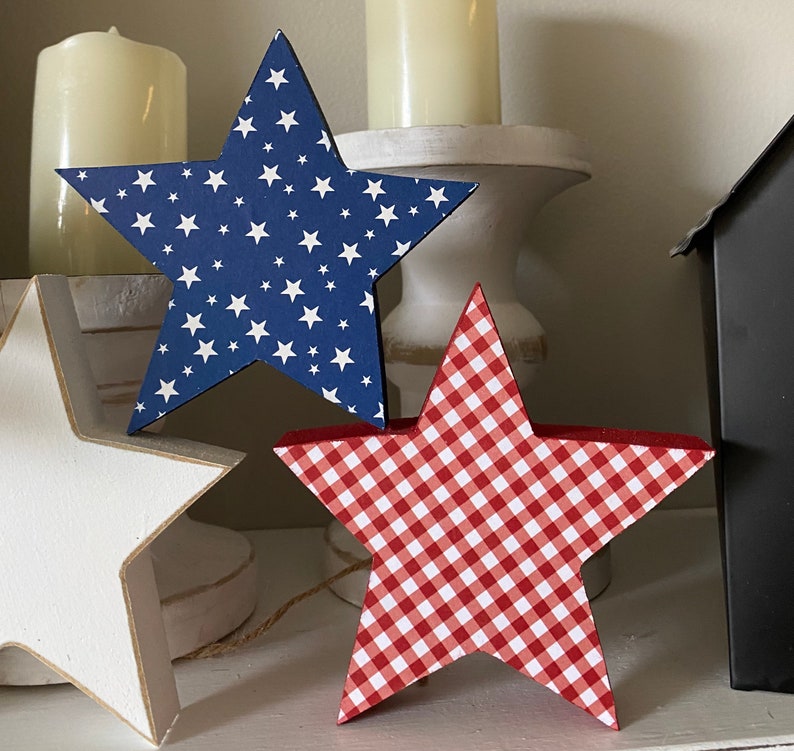 May include: Two wooden stars, one blue with white stars and one red with white gingham pattern. The stars are sitting on a white surface.