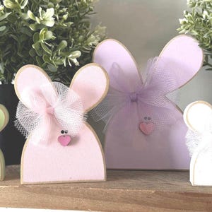 May include: Four wooden bunny figurines in pastel colors, each with a tulle bow and heart accent. The bunnies are in shades of green, pink, lavender, and white, and are arranged on a wooden shelf. Decorative Easter theme.