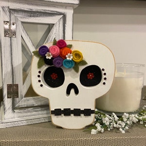 May include: White wooden sugar skull with a colourful felt flower crown. The skull has black eyes with red glitter accents and black glitter around the mouth.