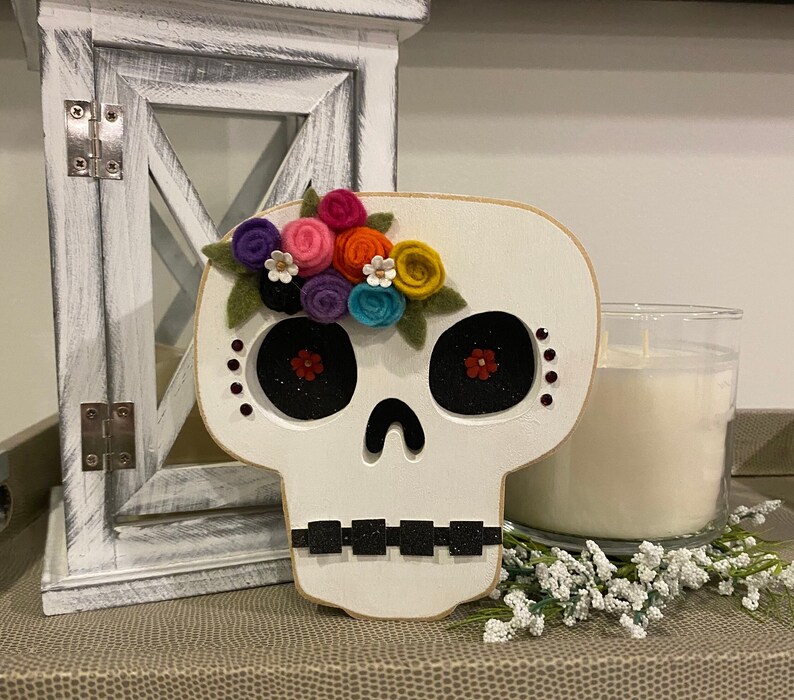 May include: White wooden sugar skull with a colourful felt flower crown. The skull has black eyes with red glitter accents and black glitter around the mouth.