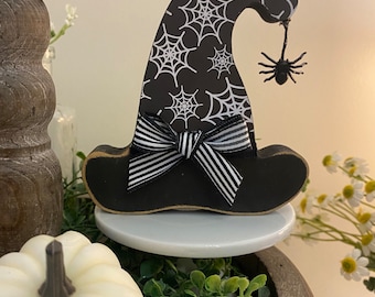 May include: Black and white witch hat with a spider on the top. The hat is decorated with a black and white striped bow. The hat is sitting on a white tiered tray.