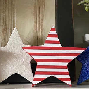 May include: Four decorative stars in red, white, and blue glitter. The stars are made of wood and have a 3D effect. They are perfect for adding a patriotic touch to your home decor.