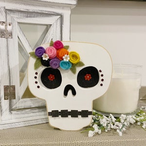 May include: White wooden sugar skull with a colourful felt flower crown. The flowers are pink, red, orange, yellow, purple, and white. The skull has black eye sockets and a black nose.