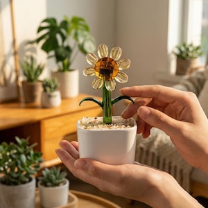 Può includere: Un girasole decorativo in vetro in un vaso quadrato bianco. Il fiore ha un centro e petali gialli, con uno stelo e foglie verdi. Il vaso è riempito di piccole pietre bianche. Un oggetto di arredamento per la casa.