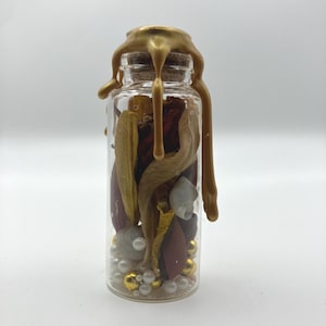 May include: A clear glass bottle sealed with a cork and topped with dripping gold wax. Inside the bottle are dried botanical elements in shades of brown and yellow, small white seashells, and a collection of white and metallic gold spherical beads.