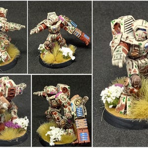 May include: A miniature sci-fi robot figurine painted in a beige, red, and green camouflage pattern. The robot features a large shoulder-mounted weapon, a blue energy vent, and stands on a circular base decorated with yellow grass and white flowers.