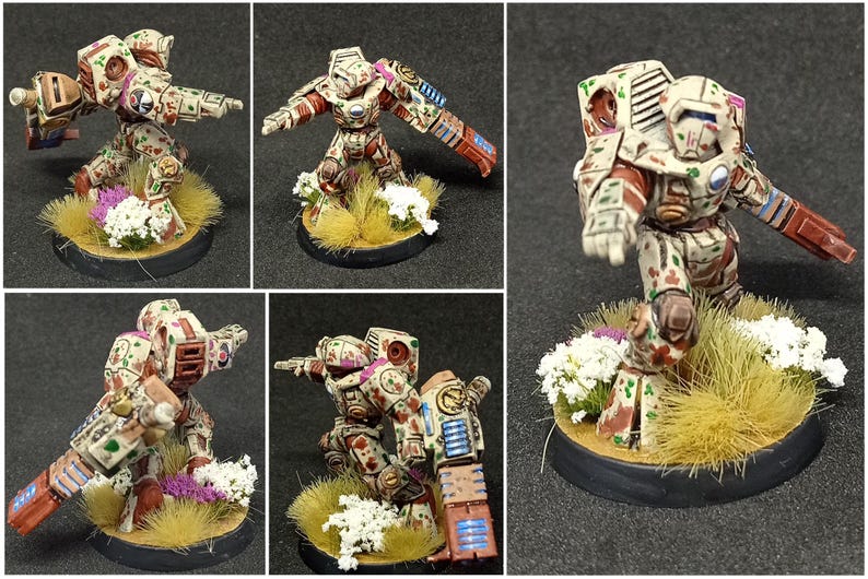May include: A miniature sci-fi robot figurine painted in a beige, red, and green camouflage pattern. The robot features a large shoulder-mounted weapon, a blue energy vent, and stands on a circular base decorated with yellow grass and white flowers.