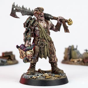 Puede incluir: Una figura en miniatura de un guerrero p&aacute;lido y musculoso con una amplia sonrisa, que lleva gafas protectoras y un taparrabos verde oliva desgarrado. La figura lleva un hacha de metal dentada sobre el hombro y sostiene una caja adornada con calaveras.