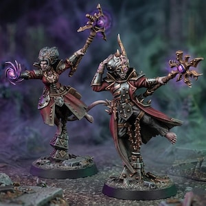 May include: Two fantasy miniature figures in red coats and dark armor standing on circular bases. One figure holds a staff with a purple orb, while the other casts a purple magical swirl. Both wear detailed metallic gear and have intricate headpieces.