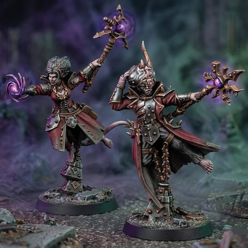 May include: Two fantasy miniature figures in red coats and dark armor standing on circular bases. One figure holds a staff with a purple orb, while the other casts a purple magical swirl. Both wear detailed metallic gear and have intricate headpieces.