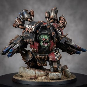 K&ouml;nnte beinhalten: Eine Miniaturfigur eines gr&uuml;nh&auml;utigen Ork-Piloten in einem schweren, verwitterten mechanischen Anzug in Schwarz und Rot. Die R&uuml;stung verf&uuml;gt &uuml;ber metallische Auspuffrohre, Schachbrettmuster und zwei am Arm montierte Kanonen auf einem Tr&uuml;mmer-Sockel.