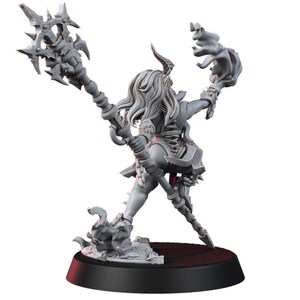 May include: A grey unpainted miniature figurine of a horned humanoid character standing on a circular base. The figure holds a large spiked staff in one hand and has flowing hair, wearing armour with a laced back detail and tattered fabric elements.