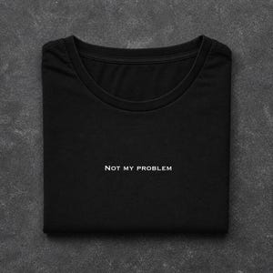 May include: Black t-shirt with the white text "NOT MY PROBLEM" across the front. The shirt is folded, revealing its crew neck. The background is a dark grey, textured surface.