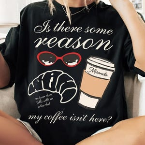 May include: Black graphic t-shirt featuring the text "Is there some reason my coffee isn't here?" with illustrations of sunglasses, a croissant, and a coffee cup. The coffee cup is labelled "Miranda".