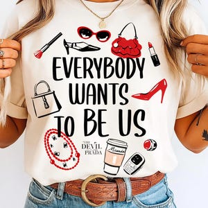 May include: A cream-coloured t-shirt with the slogan "EVERYBODY WANTS TO BE US" in black. The design includes illustrations of fashion items in red and black, such as sunglasses, a handbag, and lipstick.