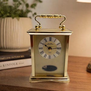 May include: A gold-colored quartz clock with a handle. The clock face has Roman numerals and the words "Seth Thomas" and "Quartz". The clock is rectangular with a handle on top.