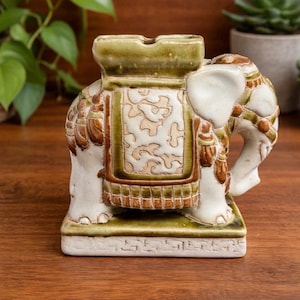May include: A ceramic elephant ashtray in white, brown, and green. The ashtray has a rectangular base and a detailed design of an elephant with a saddle. The elephant's trunk is curled, and the ashtray has a slot for cigarettes.