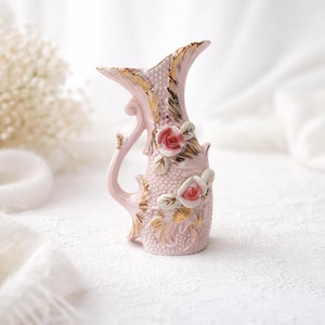 May include: A small, pink ceramic pitcher with a handle and a flared top. The pitcher is decorated with gold accents and two rose appliques. The roses are pink with green leaves. The pitcher has a textured surface.