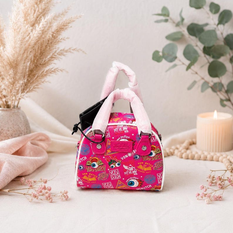May include: A pink Von Dutch handbag with white trim and pink handles. The bag features a repeating pattern of eyes, logos, and text. A black tag hangs from the handle. The bag is sitting on a white surface.