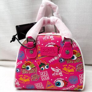 May include: A pink Von Dutch handbag with a white trim and handles. The bag is covered in various Von Dutch logos, graphics, and text. The bag has a small handle and a tag attached.
