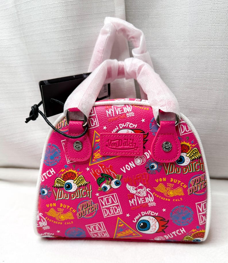 May include: A pink Von Dutch handbag with a white trim and handles. The bag is covered in various Von Dutch logos, graphics, and text. The bag has a small handle and a tag attached.