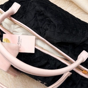 May include: A black quilted handbag with a soft velvet-like texture, featuring light pink handles and trim. The bag has a gold-toned zipper closure and a light pink rectangular tag attached that displays the word Couture in black lettering.
