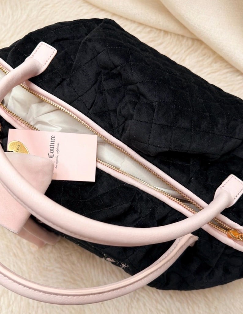 May include: A black quilted handbag with a soft velvet-like texture, featuring light pink handles and trim. The bag has a gold-toned zipper closure and a light pink rectangular tag attached that displays the word Couture in black lettering.