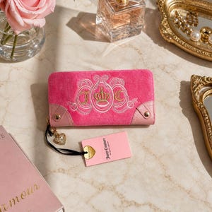 May include: A pink velour wallet with a gold embroidered crown and the letters "J" and "C". The wallet has a gold zipper pull with a heart charm and a pink tag that says "Juicy Couture".