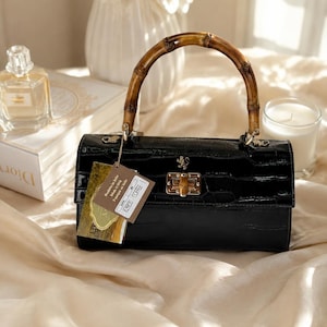 May include: A black handbag with a glossy crocodile-embossed texture, featuring a curved bamboo handle, a gold-toned metal clasp with a bamboo accent, and a small gold lion emblem. A rectangular paper tag with the text K2880 COFFEE is attached.