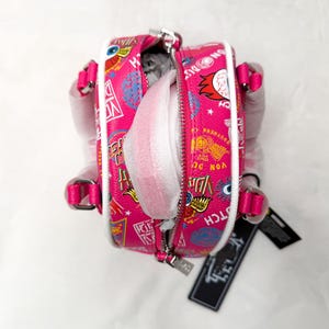 May include: A pink mini backpack with a white trim and a zipper closure. The bag is covered in colorful graphic designs and text. The straps and handle are pink. The bag is open, revealing white packing material inside.