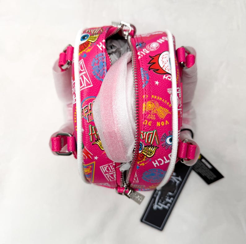 May include: A pink mini backpack with a white trim and a zipper closure. The bag is covered in colorful graphic designs and text. The straps and handle are pink. The bag is open, revealing white packing material inside.