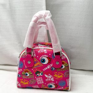 May include: A pink Von Dutch handbag with a white trim and handles. The bag is covered in a repeating pattern of eyes, logos, and text. The bag has a top handle and a zipper closure.