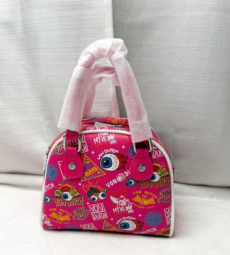 May include: A pink Von Dutch handbag with a white trim and handles. The bag is covered in a repeating pattern of eyes, logos, and text. The bag has a top handle and a zipper closure.