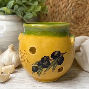 May include: A round yellow ceramic garlic keeper with a green rim and two small side handles. The front features a hand-painted illustration of a dark olive branch with green leaves and the cursive text "Ail" written in brown on the left side.