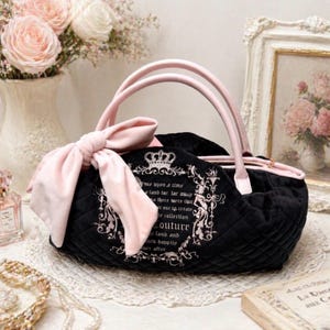 May include: A black quilted handbag featuring a large light pink bow on the side, light pink handles, and a zipper closure. The front displays a white printed design with a crown icon and text that reads "once upon a time" and "Couture".