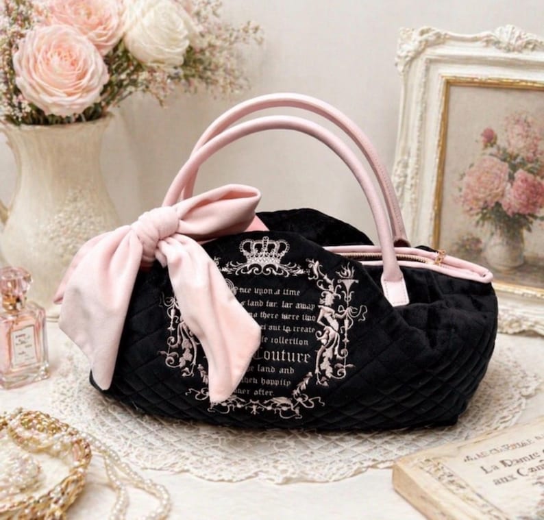 May include: A black quilted handbag featuring a large light pink bow on the side, light pink handles, and a zipper closure. The front displays a white printed design with a crown icon and text that reads "once upon a time" and "Couture".