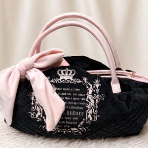 May include: A black quilted handbag featuring a large light pink bow on the side and light pink handles. The front displays a white embroidered crown and text that reads, "once upon a time a land far, far away..." with ornate decorative borders.