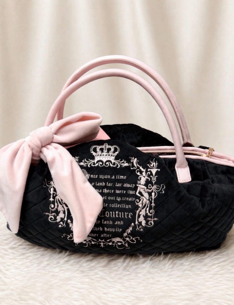 May include: A black quilted handbag featuring a large light pink bow on the side and light pink handles. The front displays a white embroidered crown and text that reads, "once upon a time a land far, far away..." with ornate decorative borders.