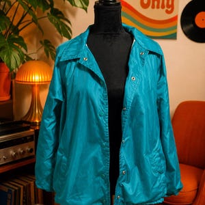 May include: Teal-colored jacket displayed on a black mannequin. The jacket has a collared neckline, snap closures, and a drawstring hem. The background includes a retro poster with the text "Groovy Vibes Only."