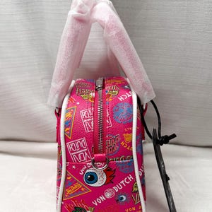 May include: Pink Von Dutch handbag with a white trim and zipper. The bag features a variety of colorful graphics and text, including the Von Dutch logo, eyes, and other designs. The bag has a top handle and a black tag.