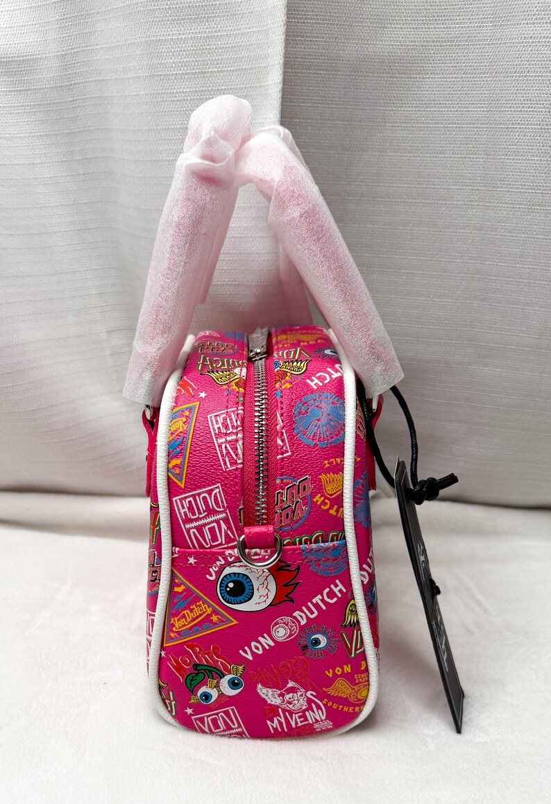 May include: Pink Von Dutch handbag with a white trim and zipper. The bag features a variety of colorful graphics and text, including the Von Dutch logo, eyes, and other designs. The bag has a top handle and a black tag.