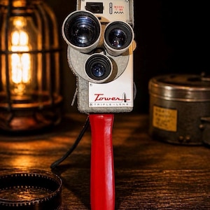 May include: Vintage Tower 8mm triple-lens movie camera with a bright red handle. The camera is silver and white with three lenses and the text "Tower" and "Triple-Lens" on the front. The camera is a retro film photography item.